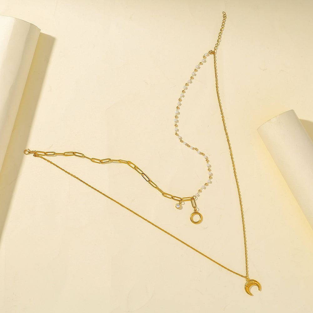 A gold-toned layered necklace set displayed on a cream-colored background with decorative paper rolls. The set includes a delicate chain with a crescent moon pendant, a paperclip chain with a small ring charm, and a beaded chain with a small clear stone accent.