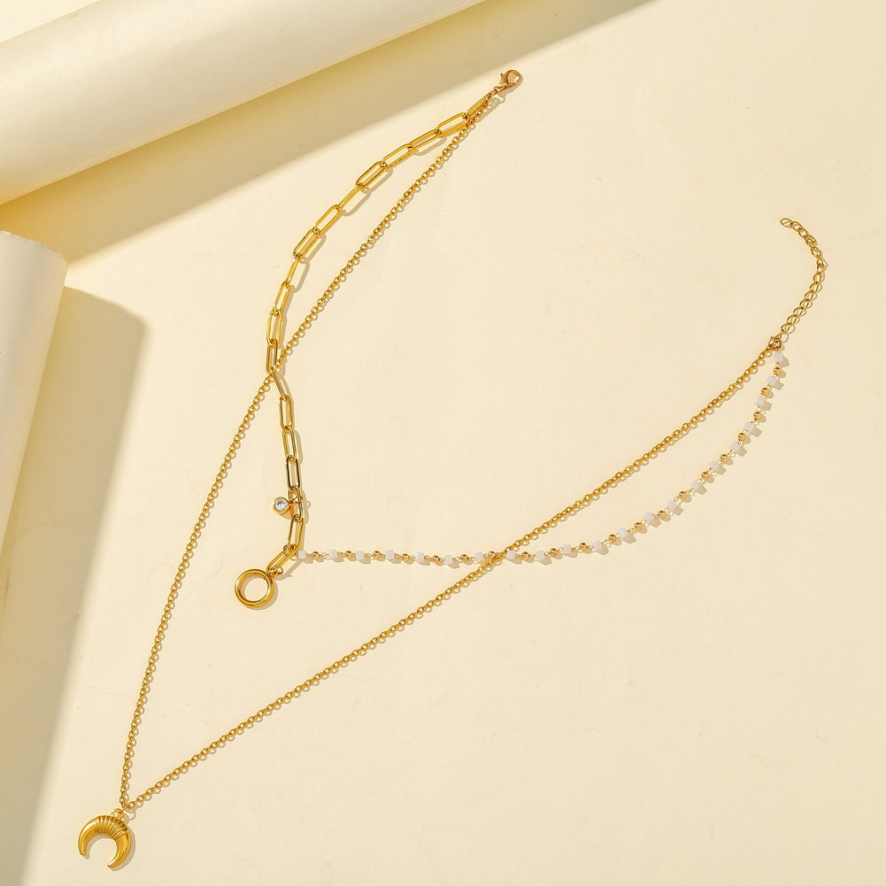 A layered gold necklace set displayed on a warm cream-colored background. The set includes a delicate chain with a small crescent moon pendant, a longer chain with a small ring pendant, and a paperclip chain with a small clear stone accent.