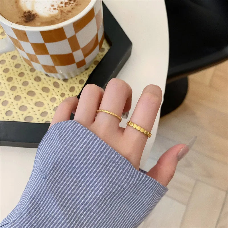 A close-up of a person's hand wearing two delicate gold rings, with a blue-and-white striped shirt sleeve visible. A checkered coffee mug and a black tray sit in the background on a light-colored table.